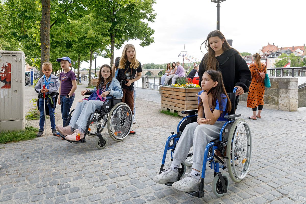 Fotografie - Kinder aus dem Kinderbeirat im Rollstuhl sitzend oder mit Langstock werden von anderen Mitgliedern des Kinderbeirates begleitet