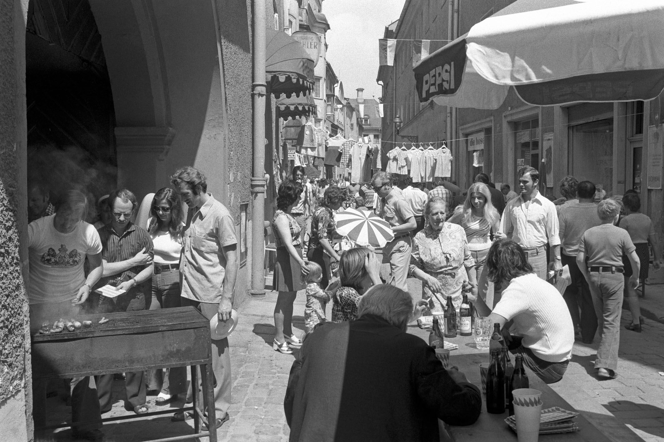 Fotografie: Grillstand, Bierbänke und Besucher in einer Gasse