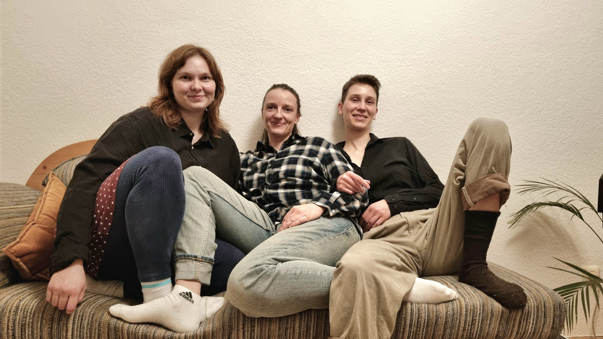 Auf dem Bild sind drei weiblich gelesene Personen zu sehen, die auf einer Couch sitzen und freundlich in die Kamera lächeln. 