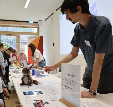 Vom rechten unteren Bildrand zieht sich ein langer Tisch bis zum oberen linken Bildrand. Dahinter steht ein Mann und eine Frau, die sich über den Tisch mit interessierten Kindern über das Thema Sensorik unterhalten.