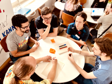 Verschiedene Menschen sitzen um einen Tisch versammelt und diskutieren miteinander. Im Hintergrund erkennt man ähnliche Gruppen um einen Tisch versammelt.