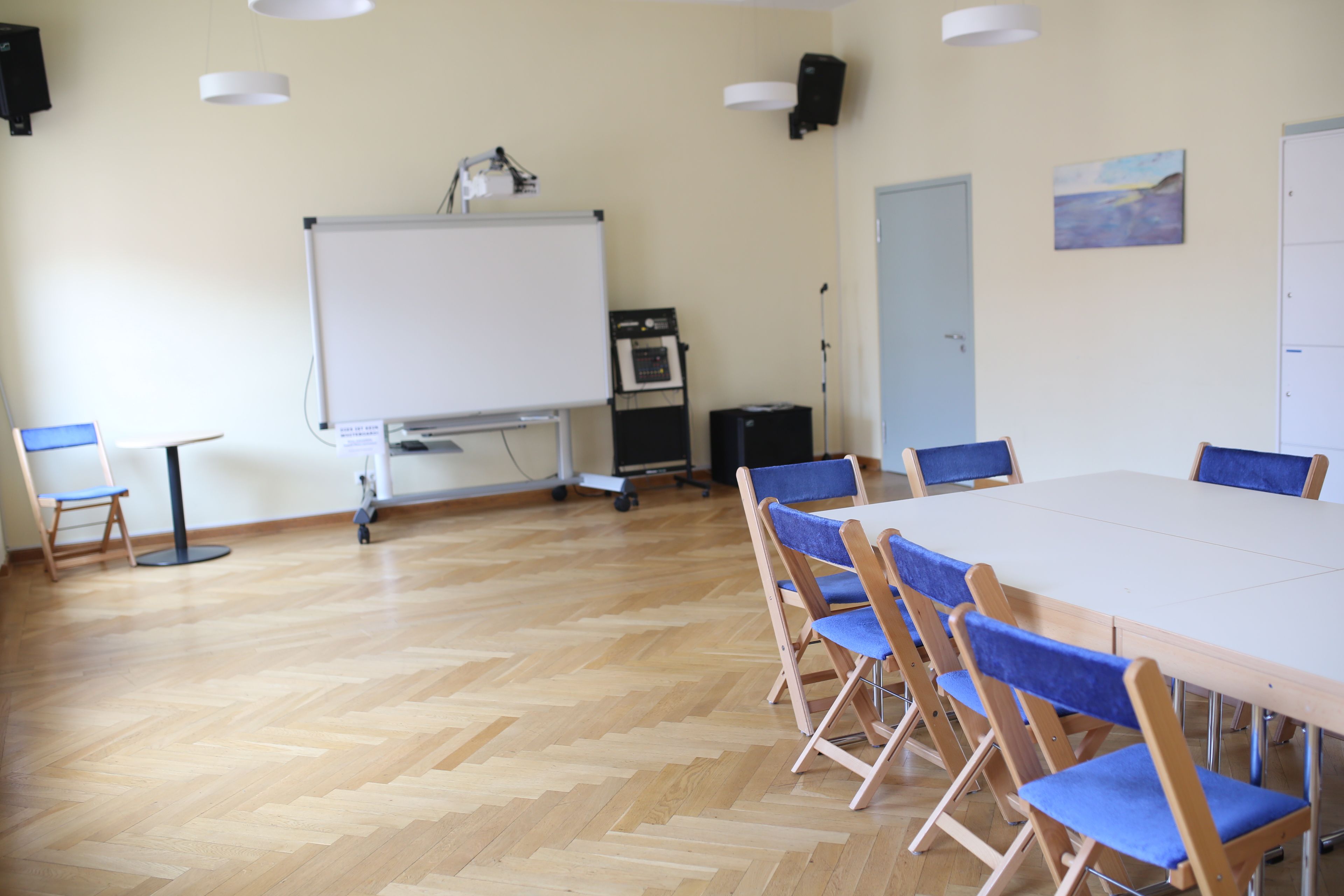 Heller Raum mit Parkett und Präsentationsleinwand mit Beamer im Hintergrund. Vorne rechts mit Stühlen und Tischen