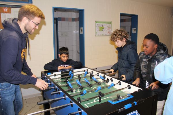 Auf dem Bild sieht man einen Tischkicker mit blauen und schwarzen Spielerfiguren. Das Spielfeld ist grün und das äußere des Kickers ist ebenfalls in blau und schwarz gehalten. Um den Kicker stehen 3 Kinder und ein Mitarbeiter des Jugendzentrums und spielen. Sie lachen und haben sichtlich Spaß.