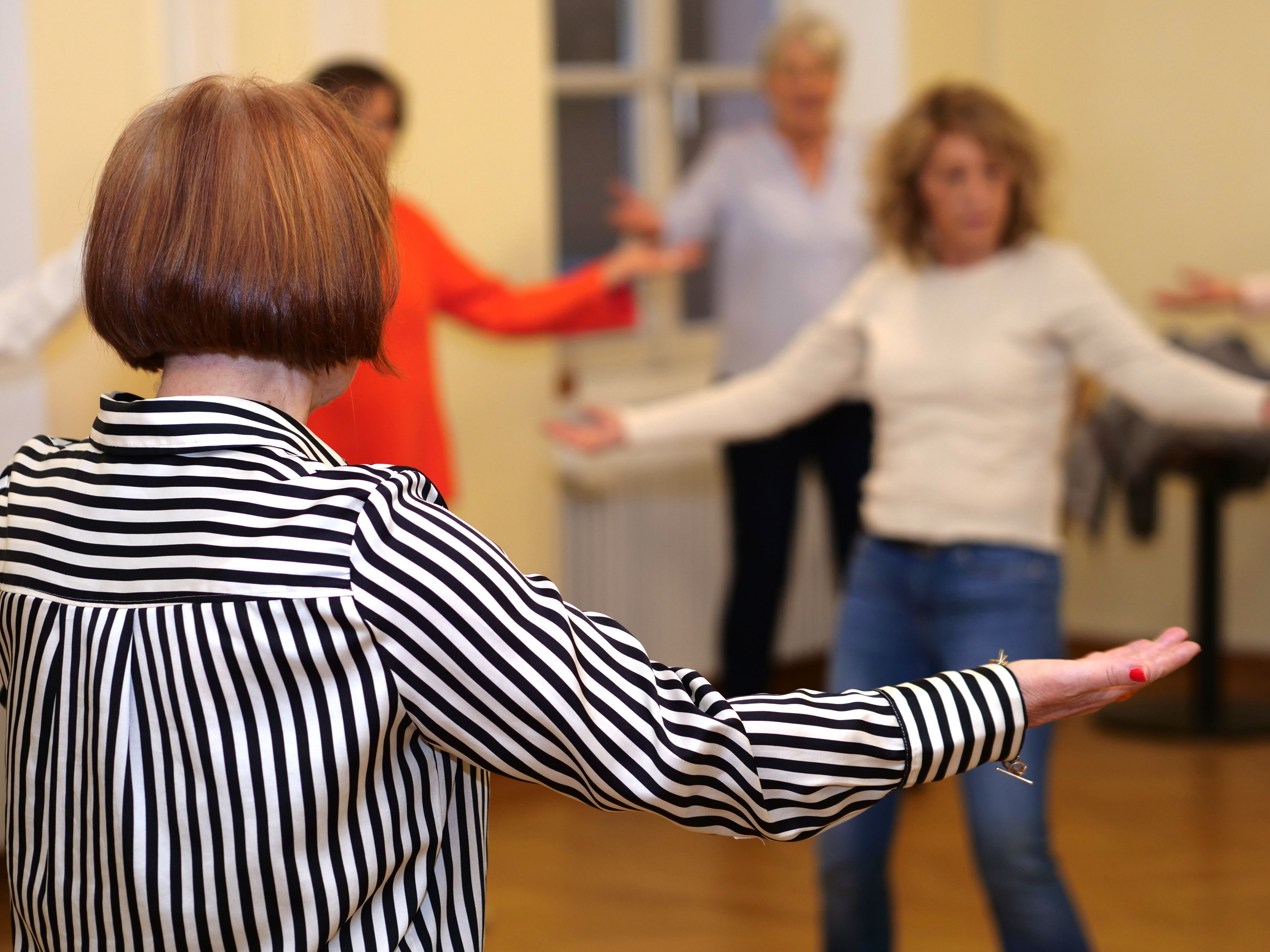 Mehrere Personen diversen Alters stehen in QiGong Pose mit ausgebreiteten Armen im Raum