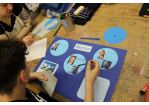 Jugendlicher sitzt mit ehrenamtlicher Betreuerin im Werkraum und erarbeitet ein Plakat für ein Referat in der Schule. Das Plakat ist blau und das Thema ist der Film Avater von James Cameron.