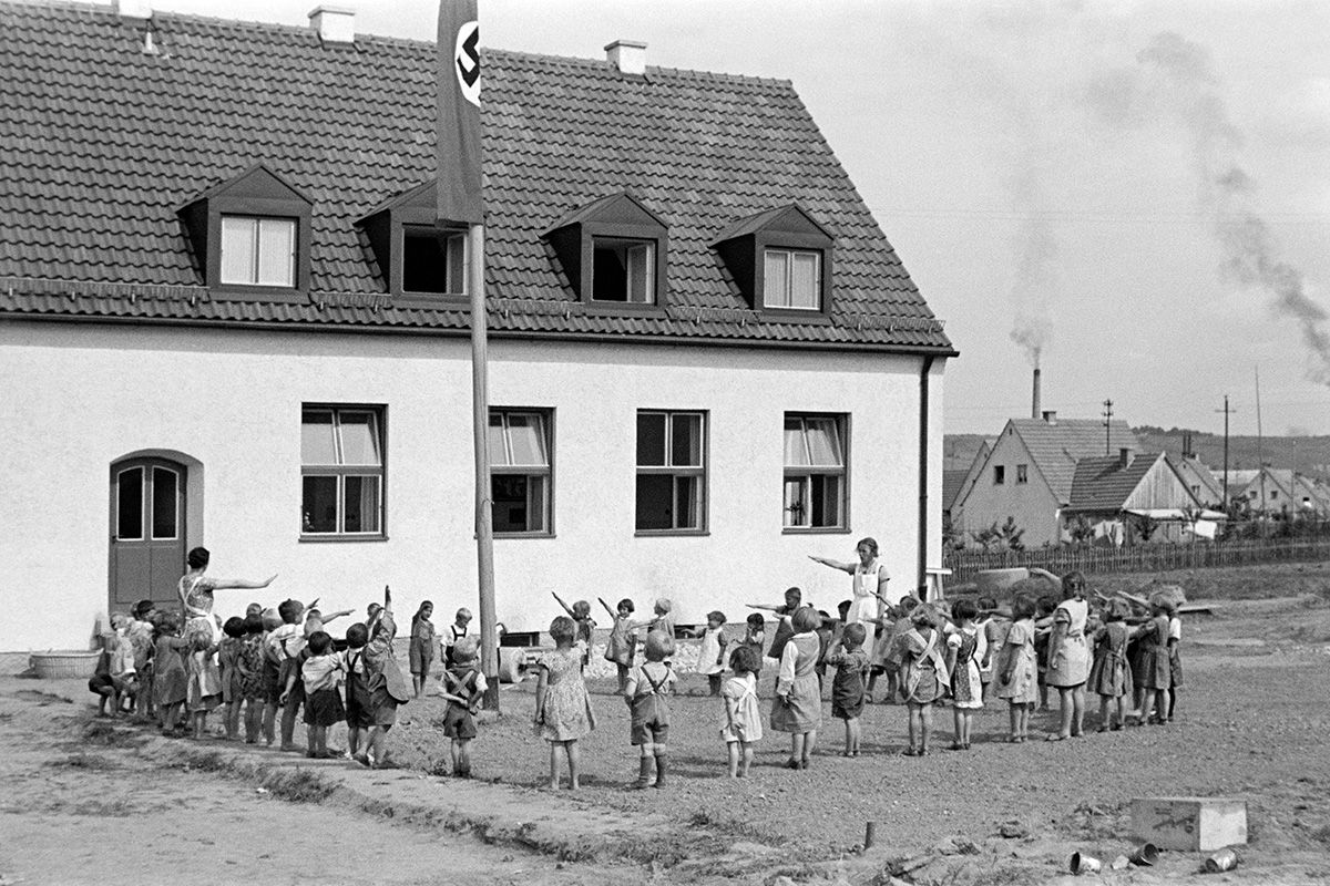 Zu sehen ist eine Kindergartengruppe mit zwei Erzieherinnen, die im Kreis um einen Fahnenmast stehen und einen Hitlergruß zeigen. Am Fahnenmast hängt eine Naziflagge. Im Hintergrund ist das Kindergartenhaus zu sehen.          