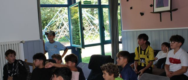 Das Bild wurde in einem Innenraum aufgenommen. Im Hintergrund sieht man ein Fenster. Im Vordergrund sind einige Kinder zu sehen, die gebannt nach links vorne gucken, wo sich der Fernseher befindet, auf dem gerade das Playstation-Spiel Fifa gespielt wird.