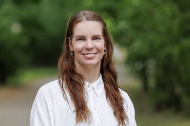 Zu sehen ist eine junge Frau in einer weißen Bluse und roten Haaren, die in die Kamera lächelt.