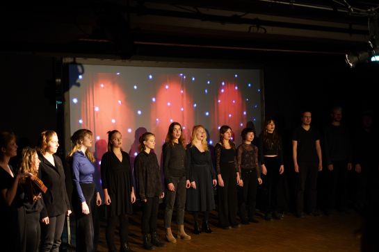 Eine Gruppe singender Personen, angeleitet von Xenia Twarz, im Theatersaal des W1 - Zentrum für junge Kultur der Stadt Regensburg.