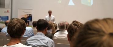 Auf dem Bild ist ein Mann zu sehen, der einen Vortrag hält. Vor dem Mann sitzen Zuschauer. Im Hintergrund ist eine große Leinwand zu sehen.