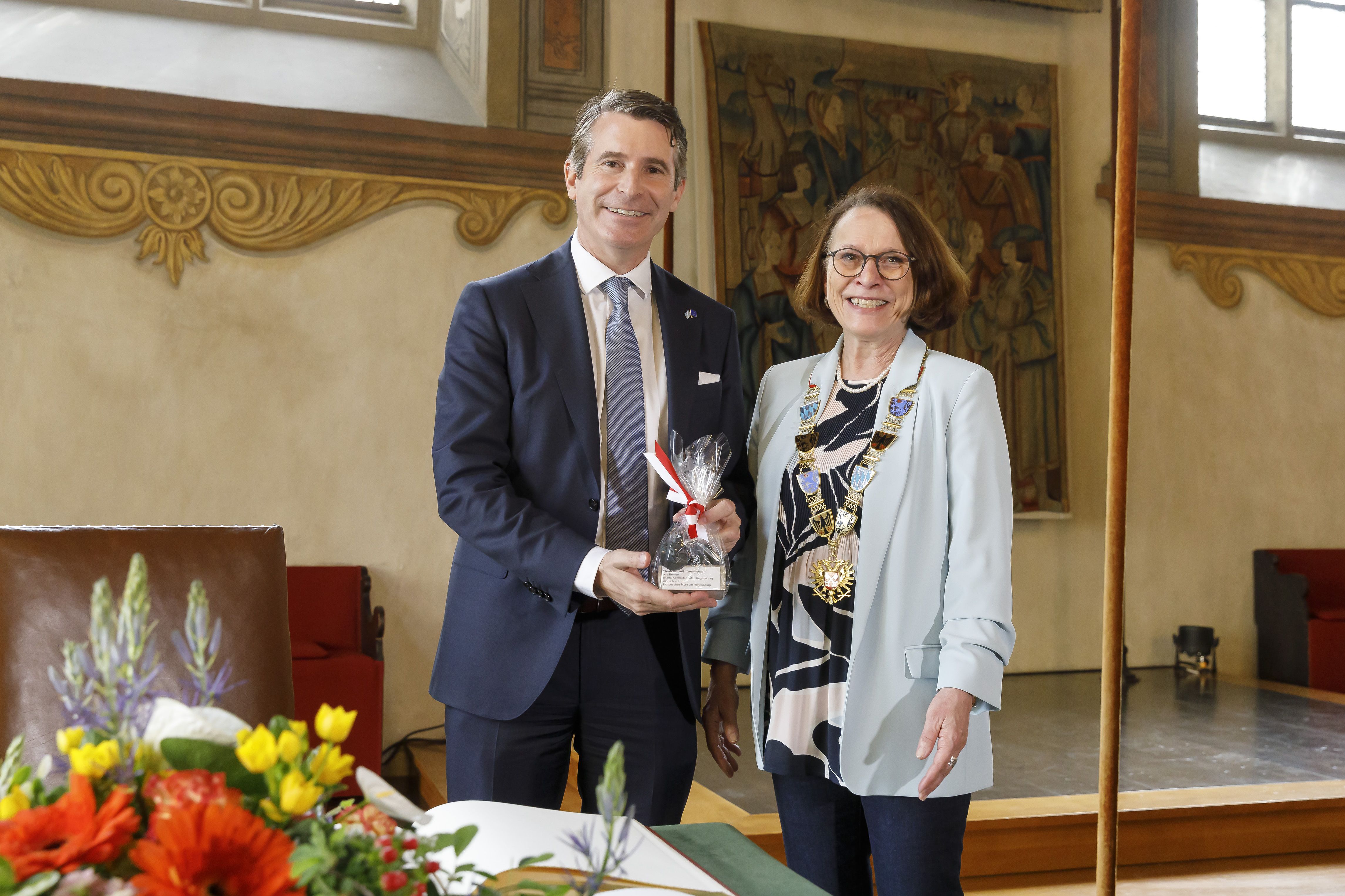Auf der Bühne im Historischen Reichssaal des Alten Rathauses hält Eric Beißwenger, Staatsminister für Europaangelegenheiten und Internationales, ein Geschenk von Oberbürgermeisterin Gertrud Maltz-.Schwarzfischer in den Händen. Beide stehen neben dem Tisch, wo der Gästebucheintrag kurz zuvor stattgefunden hat.