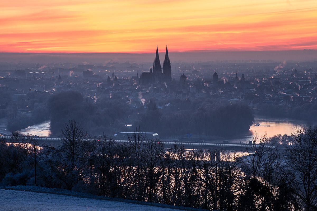Fotografie - Panoramablick auf Regensburg bei Sonnenaufgang mit Domspitzen im Morgenlicht und der Donau im Vordergrund