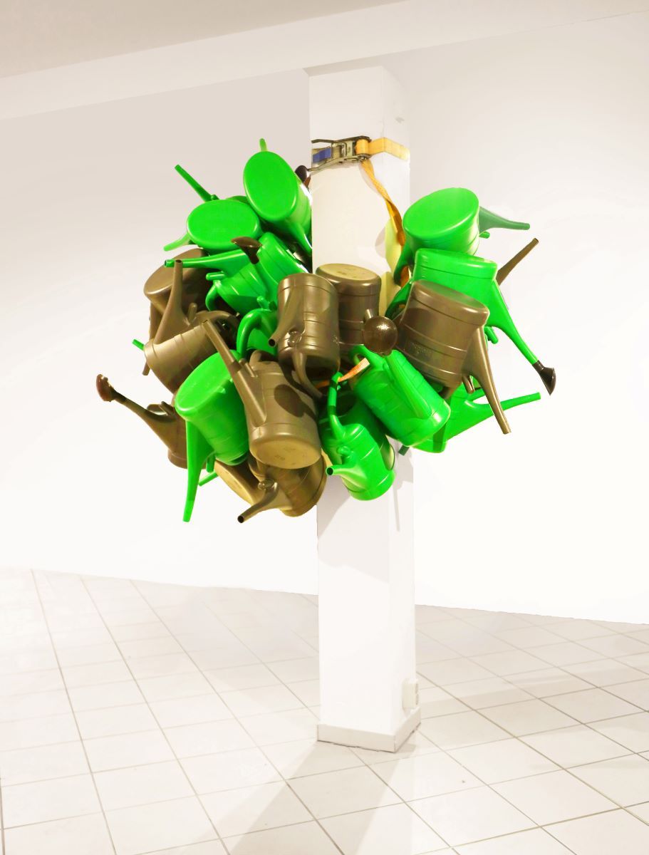 Kunstwerk: Gießkannen mit Spanngurt um Säule gewickelt