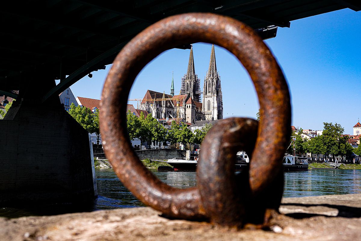 Fotografie -Blick durch eine rostige Anlegekonstruktion unter der Eisernen Brücke auf den Regensburger Dom am Donauufer.