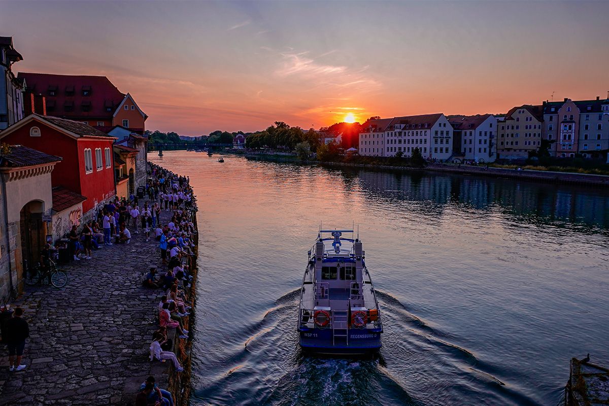 Fotografie - Donau beim Bürgerfest 2023 in Regensburg, Blick von der Steinernen Brücke, mit Boot auf dem Fluss und Menschen am Ufer bei Sonnenuntergang.