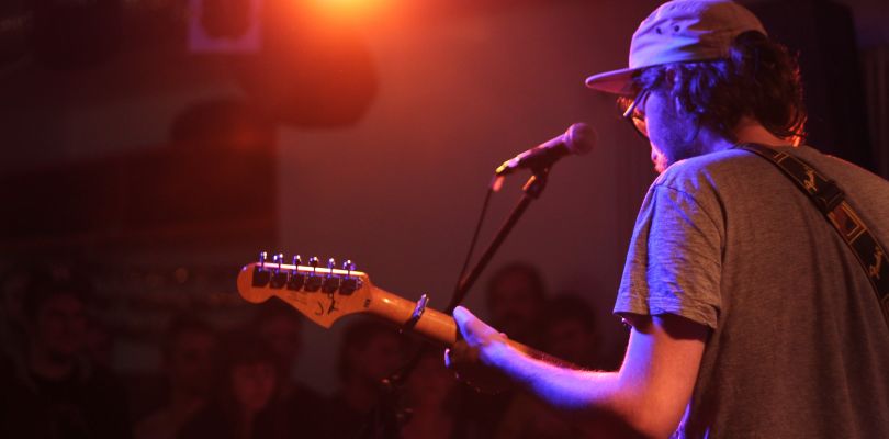 Auftritt der Band "Burkini Beach": Sänger mit Gitarre vor dem Mikrofon, Blick von hinten gegen das Licht eines Scheinwerfers.