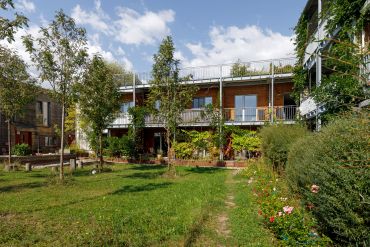 Das Foto zeigt einen Garten hinter einer Reihe miteinander verbundener Bungalows. Die Flachdachbungalows haben Balkone, an denen Fassadengrün emporwächst. Der Garten selbst ist eine Wiese mit mehreren neu gepflanzten Bäumen. Am Rand der Wiese wachsen einige Blumen und Sträucher.