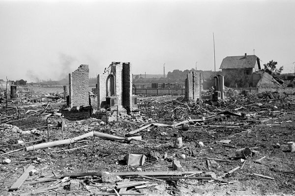 Zu sehen sind durch den Krieg zerstörte Häuser. Im Hintergrund ist ein noch intaktes Wohnhaus zu sehen. Im Vordergrund ragen Reste von Hauswänden aus Schutt und Zerstörung hervor. 