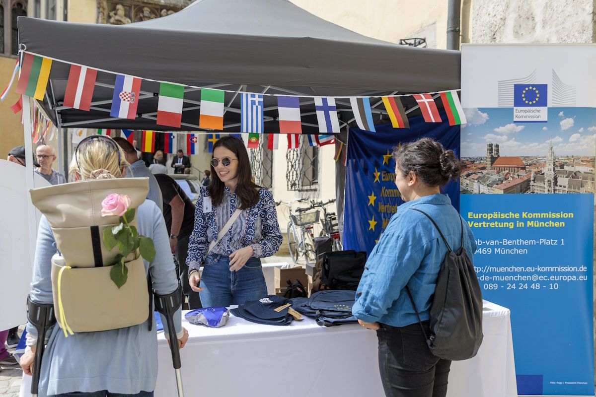 Informationsstand der Europäischen Kommission dekoriert mit kleinen Fahnen der EU-mitgliedsstaaten. Junge Frau mit Sonnenbrille spricht mit einer Besucherin. Eine weitere Besucherin beobachtet die Szene.