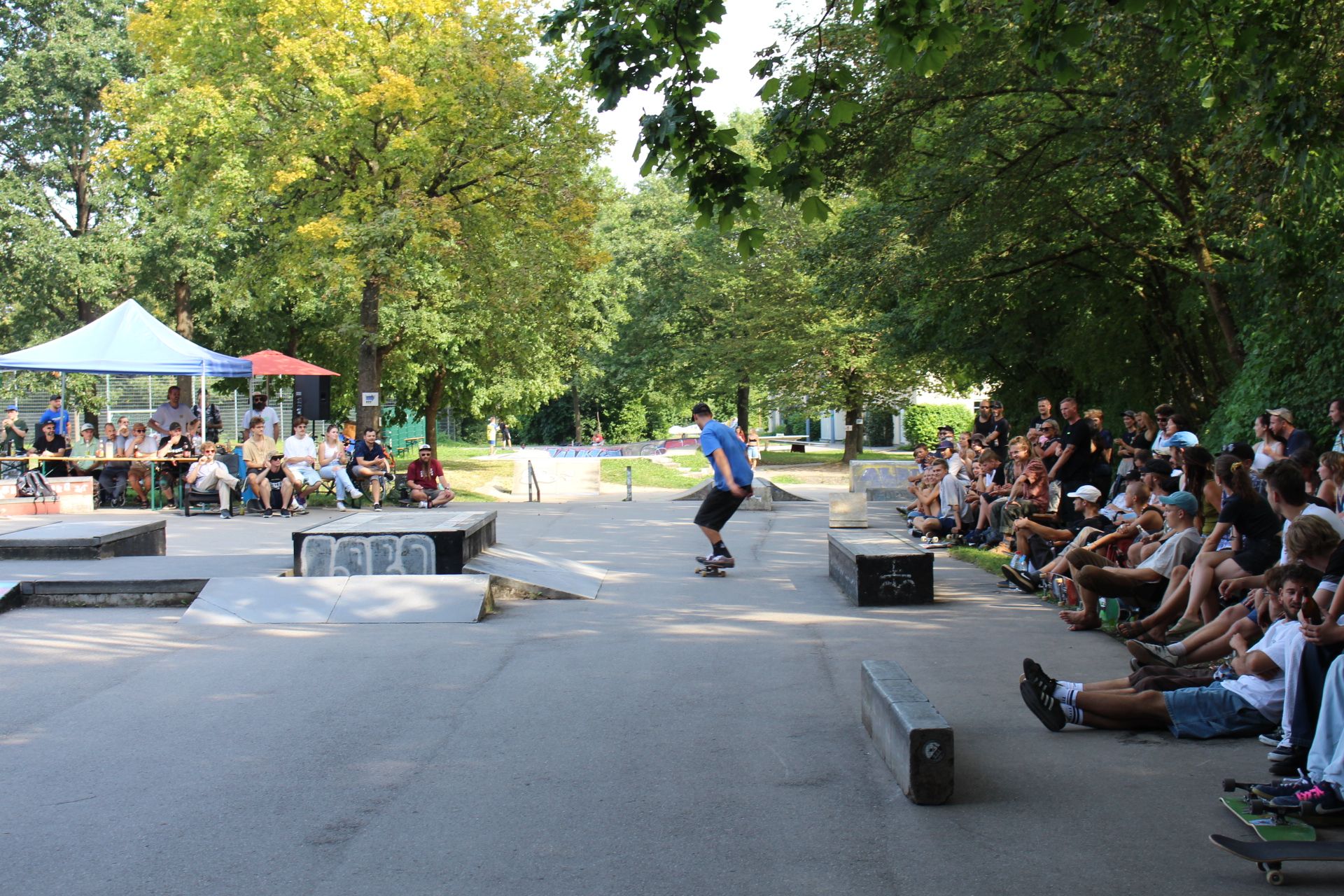 Das Foto wurde auf dem Skatepark am Jugendzentrum aufgenommen. Mittig sieht man einen Mann in blauem T-Shirt auf einem Skatepark. Rechts sitzt ein gemischtes Publikum an Zuschauenden teils auf Bänken und Sofas, teils auf Bänken. Links sieht man die weißen und blauen Pavillons der Veranstaltung. Über allem schweben die grünen Kronen der Bäume.
