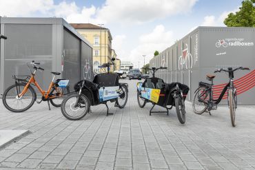 Von links zu sehen sind ein Zweirad, zwei Lastenräder und ganz rechts ein Zweirad. Im Hintergrund sind Boxen zur Unterbringung von Fahrrädern zu sehen. 