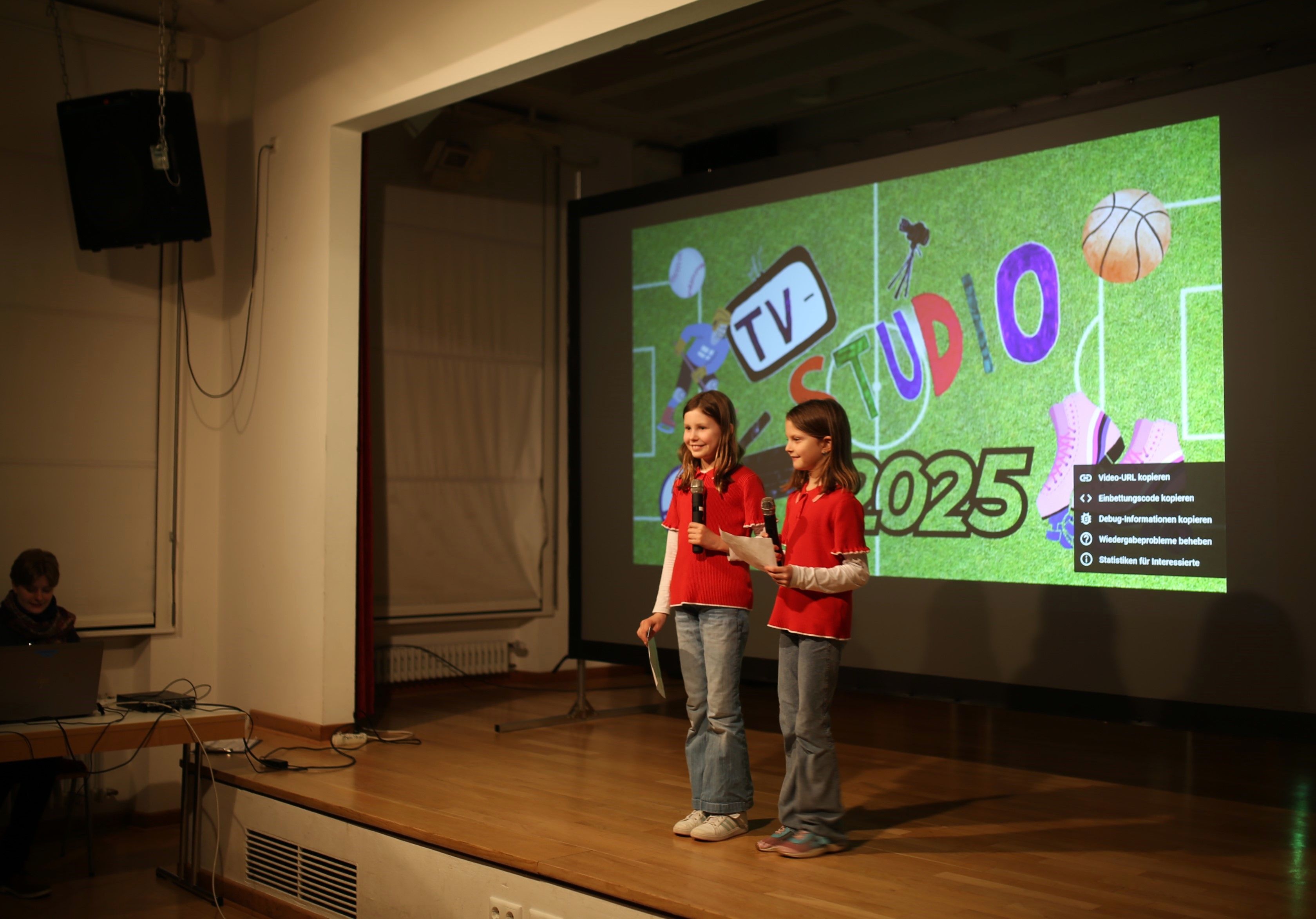 Zwei Mädchen in roten T-Shirts stehen auf einer Bühne. Sie halten Mikrofone in der Hand. Dahinter ist eine Leinwand aufgebaut. Auf dieser ist "TV-Studio 2025" zu lesen.