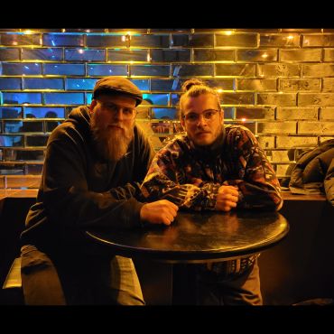 Cannibal Hector und Store1 sitzen an einem Tisch vor einer spiegelnden Backsteinwand.