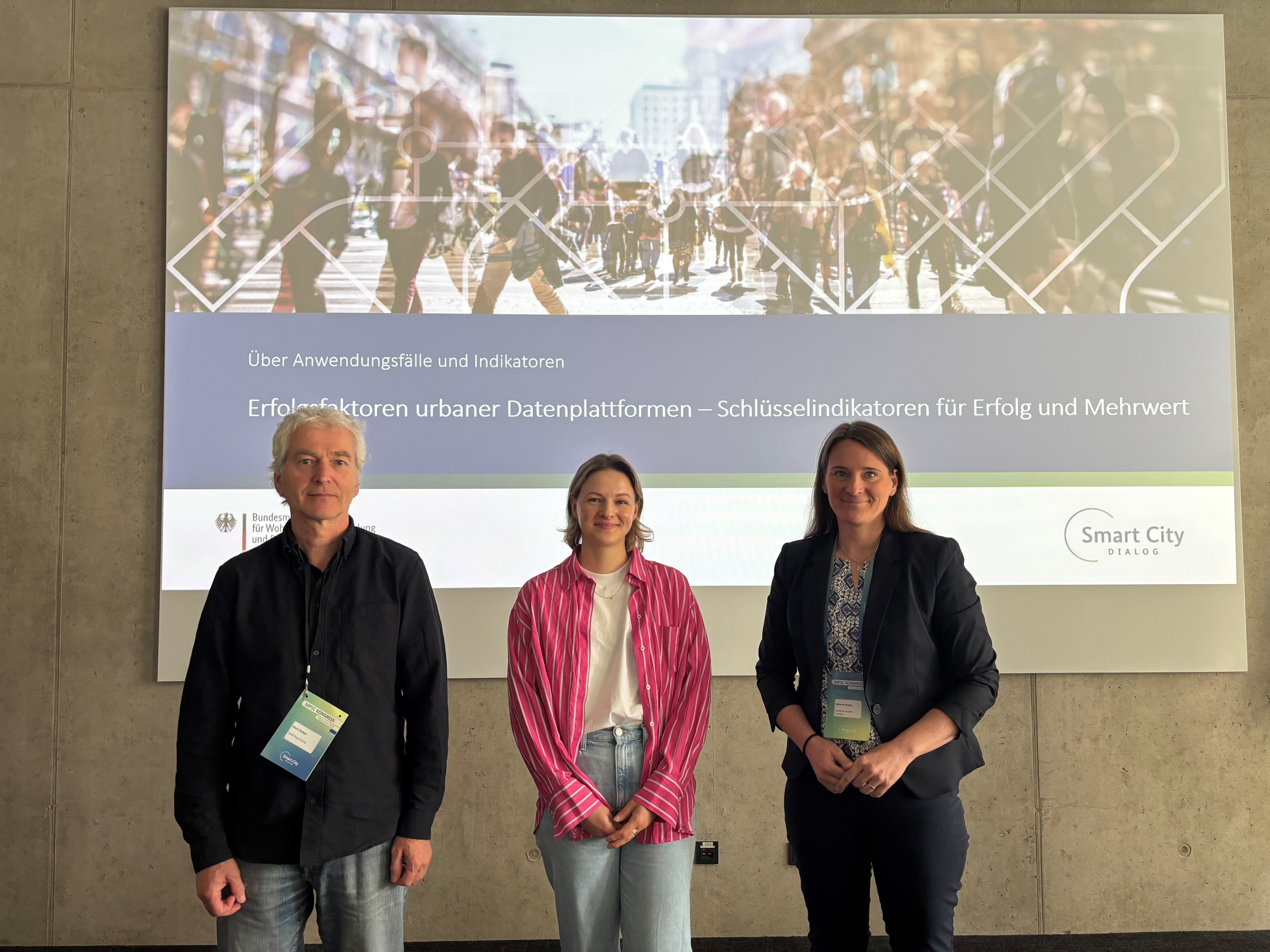 R_NEXT-Kernmitglied Mark Daibler steht mit zwei Frauen vor einer Leinwand, die eine Folie zum Thema "Erfolgsfaktoren urbaner Datenplattformen" zeigt.