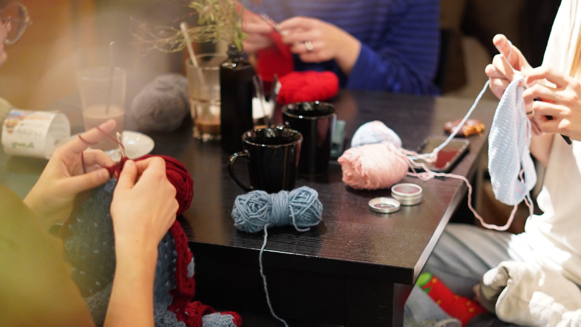 Auf dem Bild sind Hände zu sehen, die gerade Stricken und Häkeln. Es ist ein verspieltes Bokèh um das Bild herum, das die Gemütlichkeit des Nachmittages verdeutlicht. 