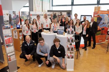Eine größere Gruppe von Personen steht und sitzt rund um einen Messestand mit Broschüren, Bannern und Präsentationselementen der Stadt Regensburg bei einer Karrieremesse im Jahnstadion.