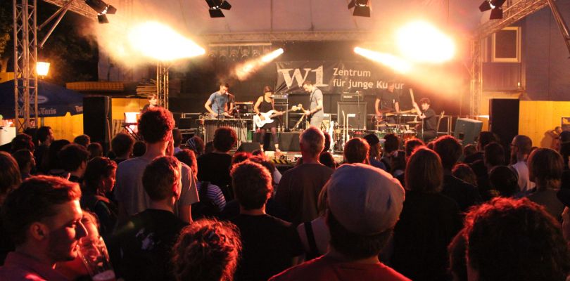 Ein Auftritt der Band "Containerhead" am Regensburger Bismarckplatz mit Blick aus dem Publikum. Im Bühnenhintergrund ein Banner mit dem Logo des W1.