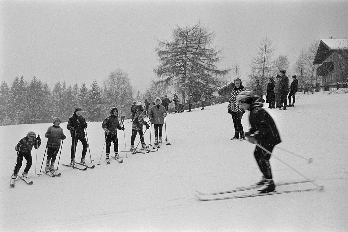 Zu sehen ist eine Winterlandschaft im Bayerischen Wald. Die Szene befindet sich an einem schneebedeckten Hügel. Im Vordergrund fährt ein Kind auf Skiern den Hang hinunter. Sechs weitere Personen stehen am Rand, ebenfalls auf Skiern und schauen zu. Im Hintergrund sind noch mehr Personen zu erkennen, allerdings ohne Skier. Auch schneebedeckte Häuser und Nadelbäume sind zu erkennen.