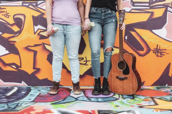 Zwei Frauen mit Spraydosen und Gitarre in der Hand vor einer besprühten Wand