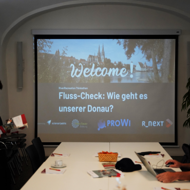In der Mitte ist die Leinwand mit der Willkommens-Folie des Vortrags erkennbar. Davor stehen weiße Tische, auf denen Blöcke und Stifte verteilt sind.