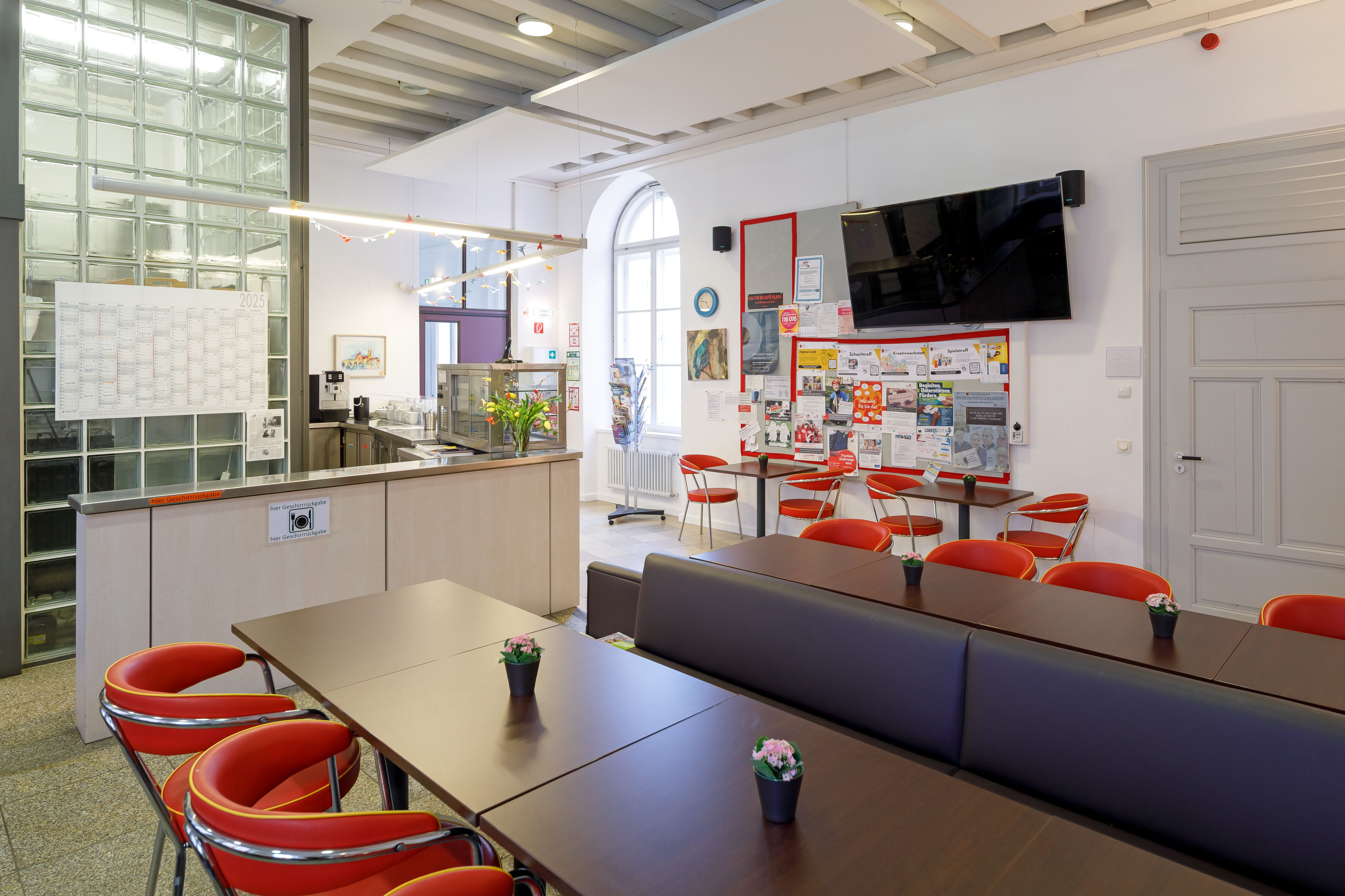 Café mit Theke links, Tischen mit Bänken und Stühlen rechts und großer Info-Pinnwand mit Bildschirm im Hintergrund. 