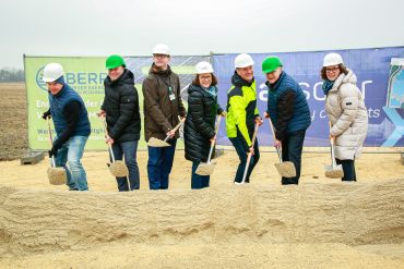 Sieben Personen stehen mit Bauhelmen vor einem Sandhaufen, in den sie symbolisch mit einem Spaten stechen. Im Hintergrund sind Bauzäune mit Bannern zu sehen.