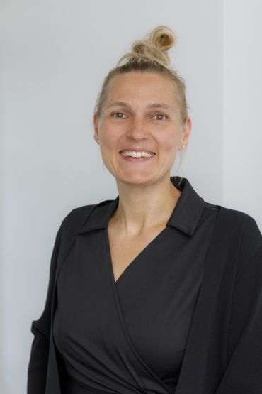 In der Mitte des Bildes steht eine breit lächelnde Frau mit blonden Haaren, die sie zu einem Dutt gebunden hat. Sie trägt Perlenohrringe und ein schwarzes Wickelkleid.