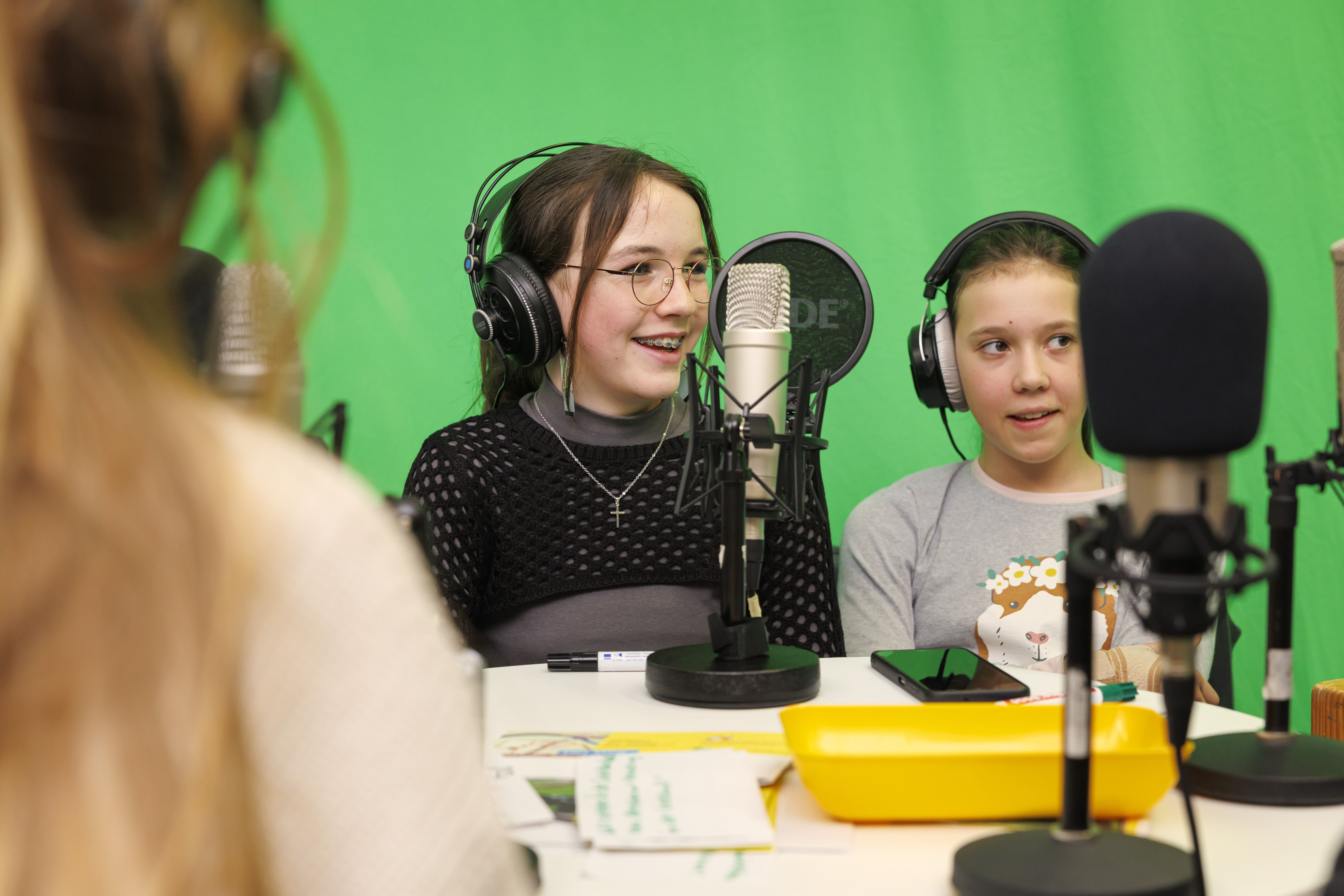 Ein Kind mit braunen Haaren und Kopfhörern sitzt vor einem Podcast-Mirkofon und strahlt. Daneben sitzt ein weiteres Mädchen mit braunen Haaren und hört zu.