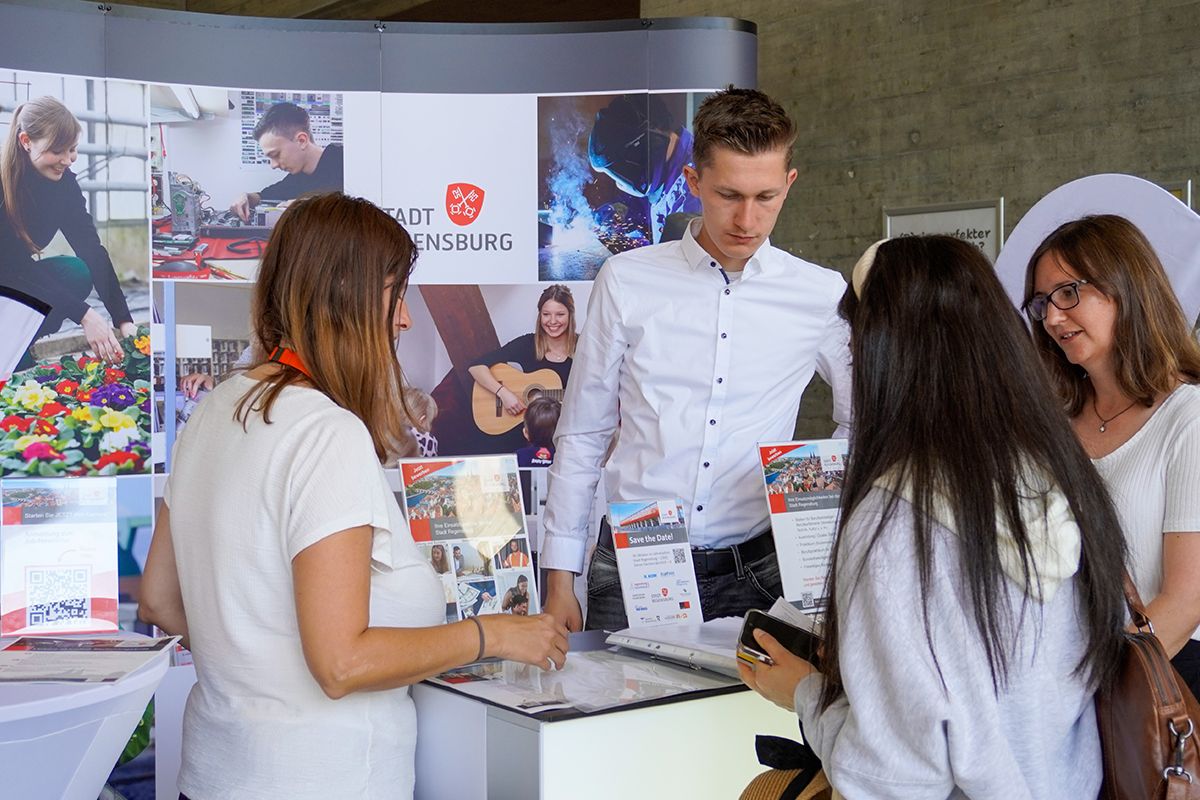 Personen führen ein Gespräch an einem Messetresen, während im Vorder- und Hintergrund Infomaterial und Banner des städtischen Karriere- und Ausbildungsangebots zu sehen sind.