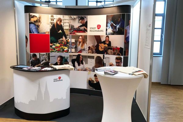 Fotografie - Messestand der Stadt Regensburg - im Vordergrund ein Stehtisch und ein Counter, im Hintergrund eine Bilderwand mit Fotos städtischer Bediensteter