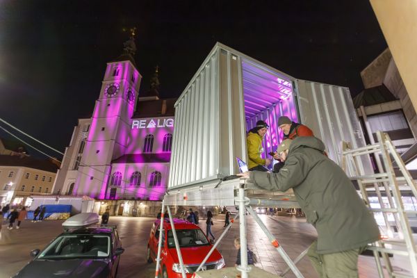 Fotografie: Drei Männer besprechen die Beleuchtung in einem Container, im Hintergrund die beleuchtete Neupfarrkirche.