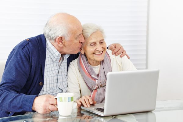 Fotografie: Ein Seniorenpaar sitzt vor einem Laptop. Der ältere Herr hat einen Arm um die Dame gelegt und gibt ihr ein Küsschen.