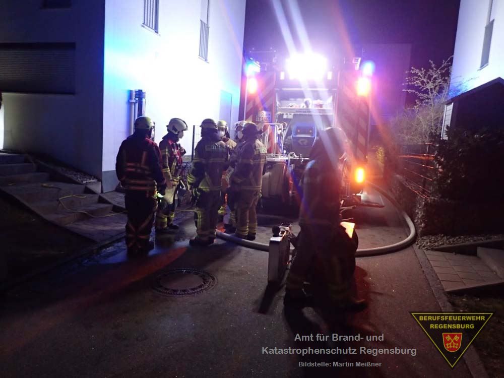 Einsatzkräfte der Feuerwehr an einer Einsatzstelle, Löschfahrzeug und Gebäude im Hintergrund.