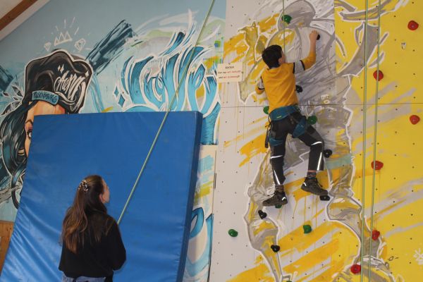 Auf dem Bild ist die Kletterwand im Jugendzentrum Arena abgebildet. Sie ist in grau und gelb bemalt und zeigt einen grauen Bergsteiger auf gelben Hintergrund. Daneben ist eine blaue Turnmatte an die Wand gelehnt. Im Vordergrund des Bildes steht eine Mitarbeiterin des Jugendzentrum mit einem schwarzen Bild und sichert mit einem grünen Seil ein Kind beim Klettern. Im Hintergrund sieht man das Kind in der Mitte der Kletterwand. Es trägt eine dunkle Hose, ein gelbes T-Shirt und den blau-grünen Klettergurt durch den es gesichert wird.