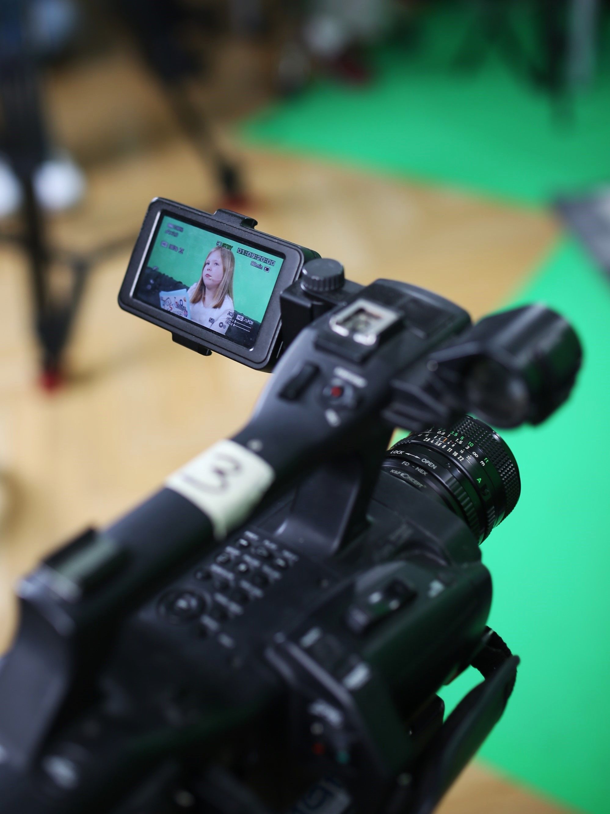Foto einer Filmkamera. Dahinter ist Greenscreen zu erkennen. Auf der Kamera ist das Vorschaubild aktiviert. Dort ist ein Mädchen mit Moderationskarten zu erkennen. 