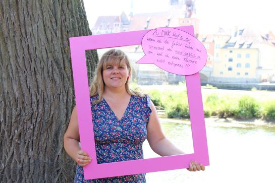 Fotografie: Michaela Schindler hält einen Bilderrahmen mit der Sprechblase „Zu PINK wird es mir, wenn ich das Gefühl haben muss, unnormal oder nicht weiblich zu sein, weil ich einem Klischee nicht entspreche!!!“. (C) Judith Pöpping