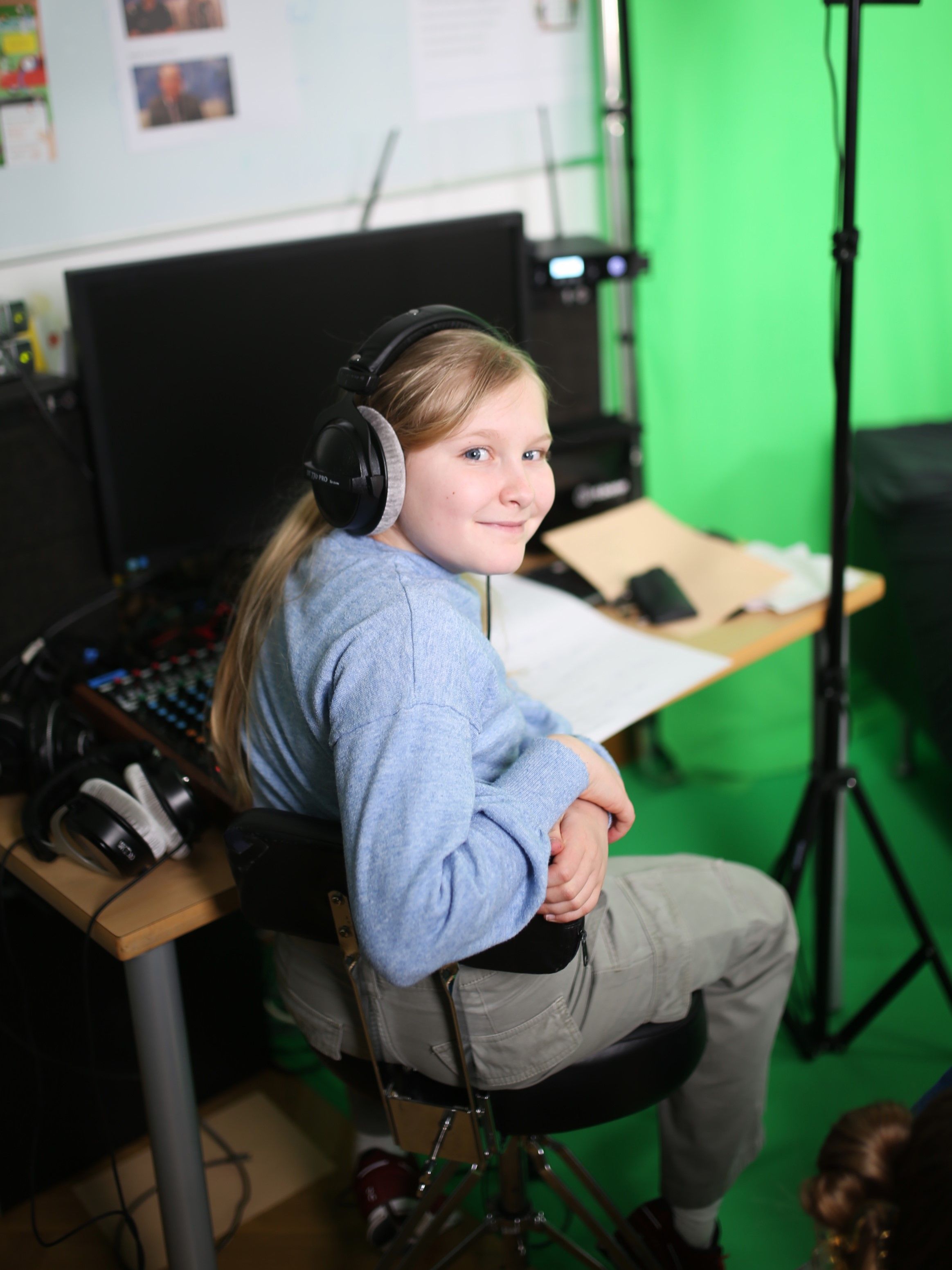 Ein Mädchen sitzt mit Kopfhörern vor einem Mischpult und einem PC und lächelt in die Kamera. Hinter der Szenerie ist ein Greenscreen zu erkennen.