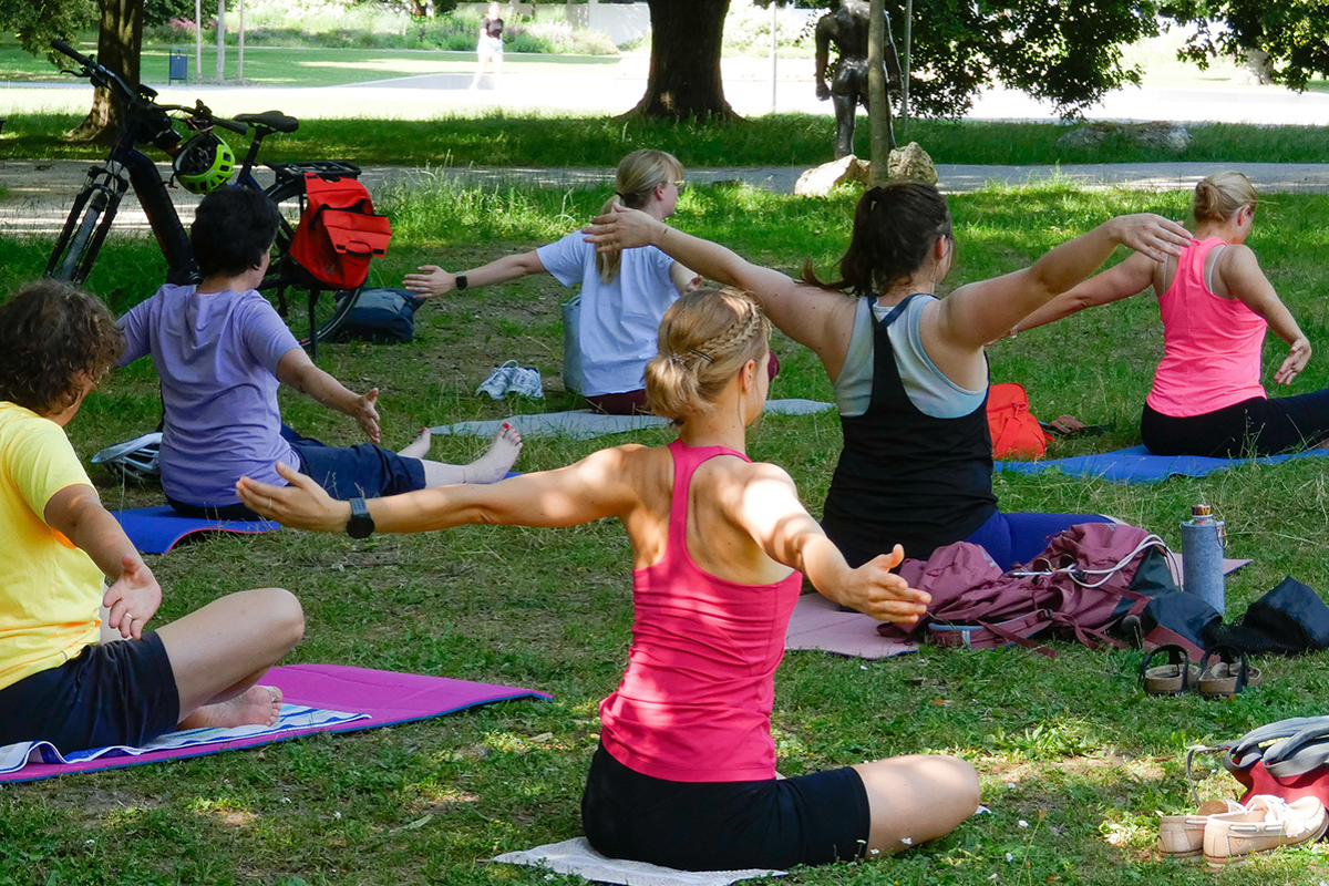 Fotografie -JMehrere junge Personen sitzen auf Yoga-Matten in einem Park in Regensburg und absolvieren eine Yoga-Übung.