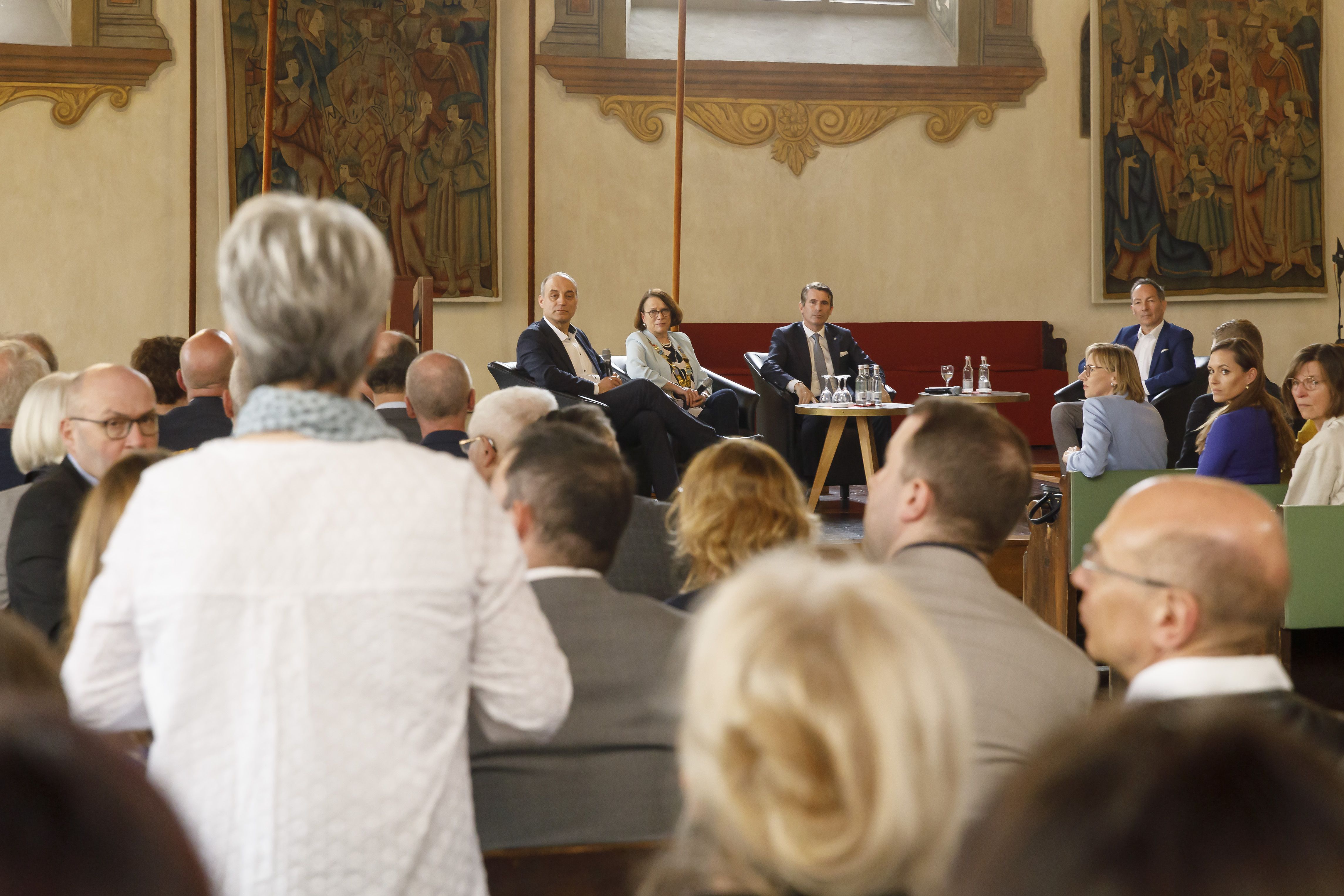  Blick vom Publikum aus im historischen Reichssaal des Alten Rathauses auf die Bühne. Eine Frau im Publikum steht. Die Augen der der vier Teilnehmer an der Podiumsdiskussion auf der Bühne sind auf sie gerichtet.