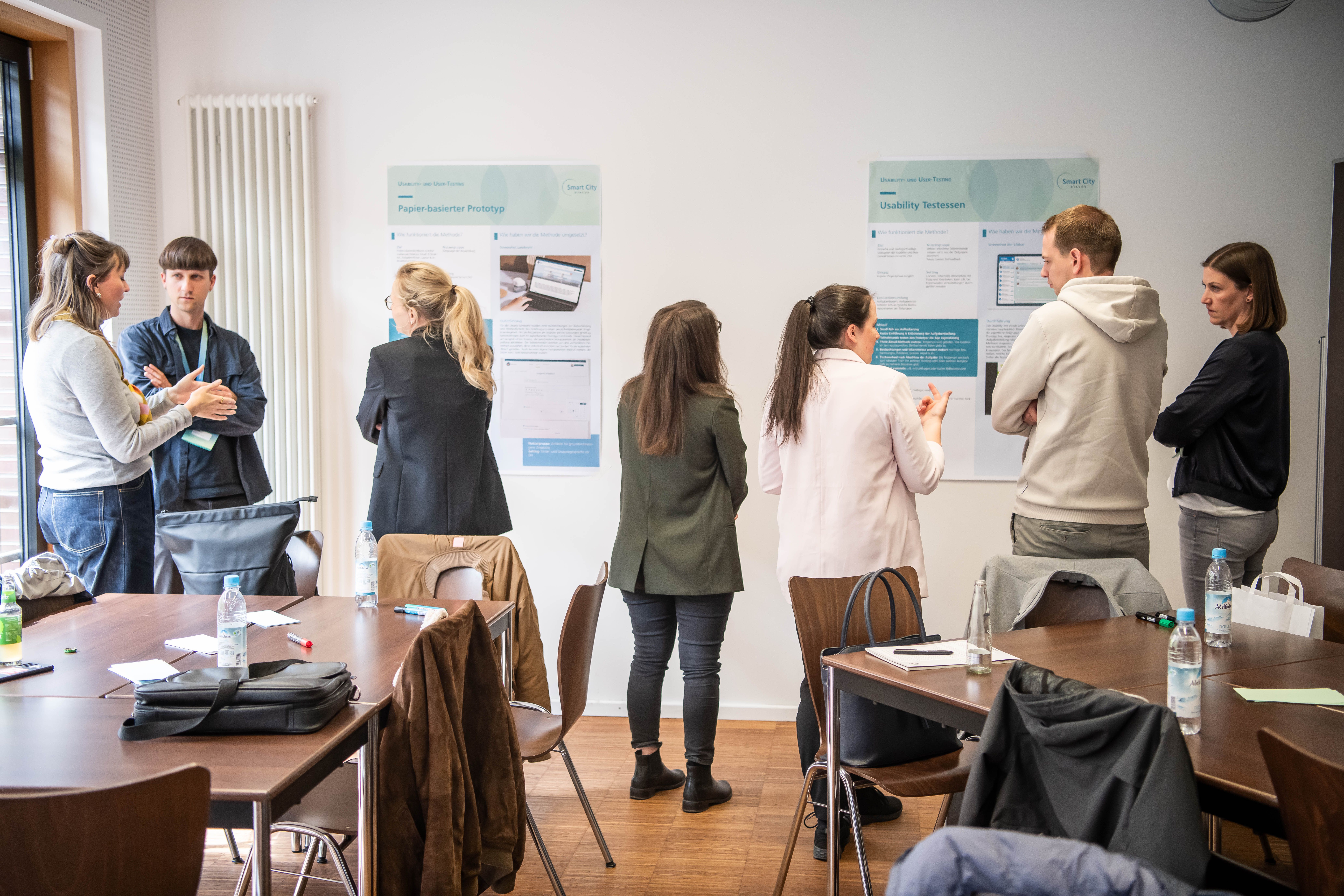 Emir Skulic steht im hinteren linken Bildrand an einer Wand und unterhält sich mit zwei Frauen. An der Wand hängen verschiedene Infografiken; vor Ihnen stehen Tische zu Gruppentischen zusammengeschoben.